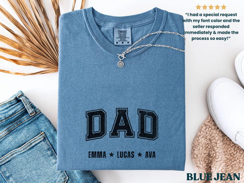 May include: A blue jean-colored t-shirt with the word "DAD" in black block letters. Below the word "DAD" are the names "EMMA", "LUCAS", and "AVA". A silver necklace with a pendant rests on the shirt. The shirt is folded and placed on a white surface.
