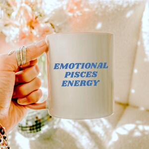 Op de afbeelding: Een witte keramische mok met een roze handvat. De mok heeft blauwe tekst met de tekst "Emotional Pisces Energy".