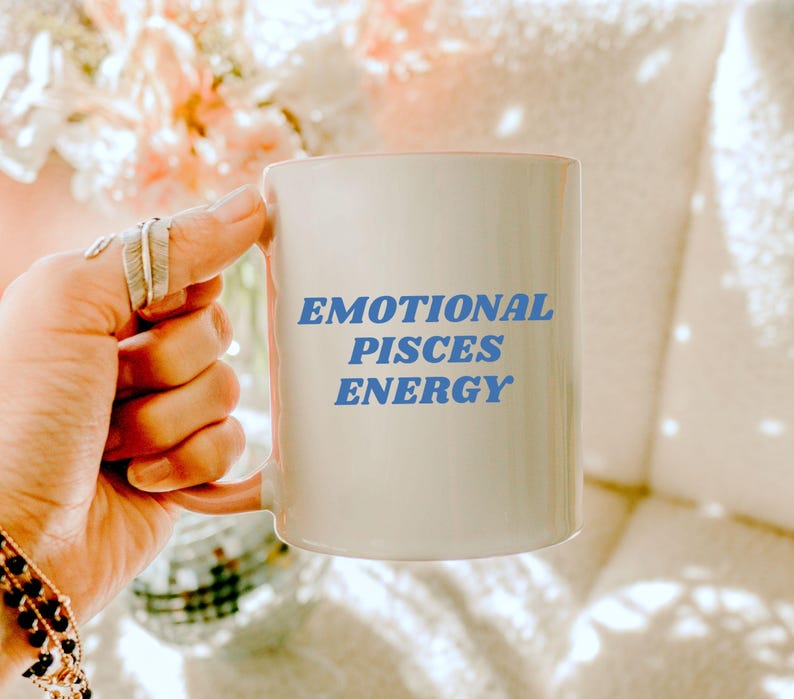 Op de afbeelding: Een witte keramische mok met een roze handvat. De mok heeft blauwe tekst met de tekst "Emotional Pisces Energy".