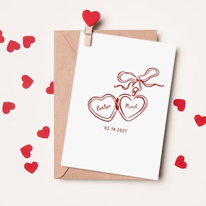 May include: A white card with a red heart-shaped locket with the names "Evelyn" and "Noah" inscribed on the locket. The date "03.14.2021" is printed below the locket.