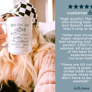 May include: White ceramic mug featuring the text "Reel Cool Dad" and green fish illustrations. The mug also has the text "ESTD 2026". A person is holding the mug, wearing a black and white checkered hat.