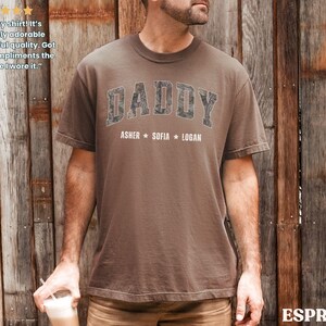 K&ouml;nnte beinhalten: Braunes T-Shirt mit dem Wort "DADDY" in einem Camouflage-Design auf der Brust, darunter die Namen Asher, Sofia und Logan. Das Shirt hat einen Rundhalsausschnitt und kurze &Auml;rmel.