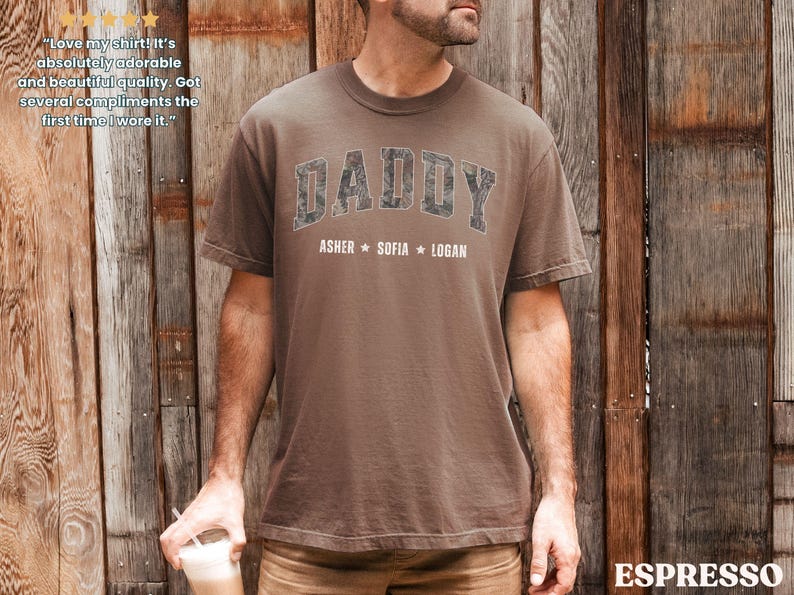 K&ouml;nnte beinhalten: Braunes T-Shirt mit dem Wort "DADDY" in einem Camouflage-Design auf der Brust, darunter die Namen Asher, Sofia und Logan. Das Shirt hat einen Rundhalsausschnitt und kurze &Auml;rmel.