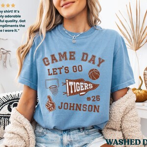 May include: Pale blue "Game Day" t-shirt with brown graphics, including a basketball, pennant, and the text "Tigers #25 Johnson." The shirt has a vintage, washed denim appearance. The shirt is a short-sleeved crew neck.