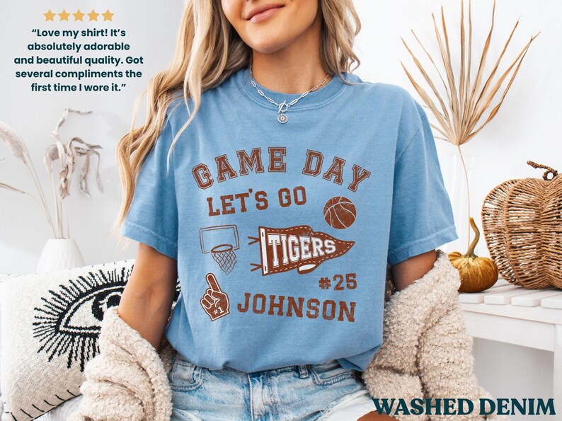 May include: Pale blue "Game Day" t-shirt with brown graphics, including a basketball, pennant, and the text "Tigers #25 Johnson." The shirt has a vintage, washed denim appearance. The shirt is a short-sleeved crew neck.