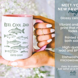 May include: White ceramic mug with a pink interior and handle, featuring the text "REEL COOL DAD" and illustrations of fish. The mug also has the text "ESTD 2026".