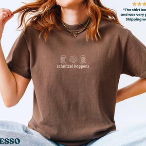 May include: Espresso-colored t-shirt with the phrase "schnitzel happens" and three small line-drawn images above the text. The shirt is a short-sleeved crew neck.