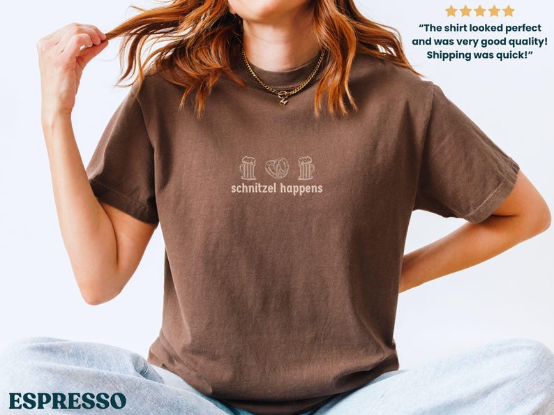 May include: Espresso-colored t-shirt with the phrase "schnitzel happens" and three small line-drawn images above the text. The shirt is a short-sleeved crew neck.
