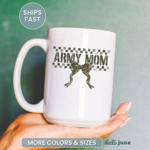May include: White ceramic mug with the text "ARMY MOM" in a distressed font, above a camouflage bow. The mug has a large handle and is held in a hand. The mug is a gift for an Army Mom.