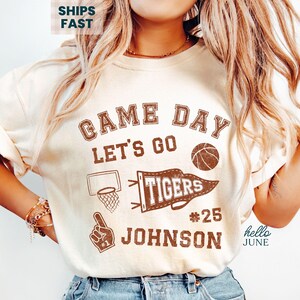 May include: Ivory t-shirt with a brown graphic design. The design includes the words "Game Day Let's Go," a basketball, a pennant that says "Tigers," a basketball hoop, a foam finger, and the name "Johnson" with the number 25.