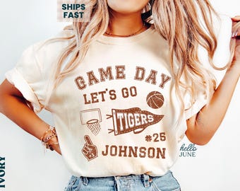 May include: Ivory-colored t-shirt with a brown graphic design. The design includes the words "Game Day Let's Go," a basketball, a pennant that says "Tigers," a basketball hoop, a foam finger, and the name "Johnson" with the number 25.