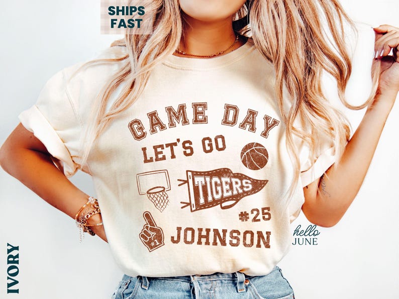 May include: Ivory t-shirt with a brown graphic design. The design includes the words "Game Day Let's Go," a basketball, a pennant that says "Tigers," a basketball hoop, a foam finger, and the name "Johnson" with the number 25.
