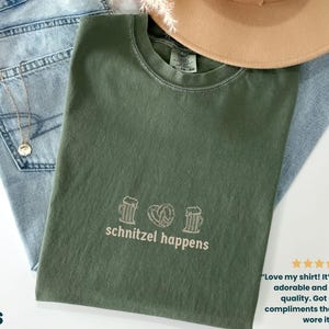 May include: An olive green t-shirt with the phrase "schnitzel happens" printed in white, along with beer mugs and a pretzel graphic. The shirt is laid on top of blue jeans and a tan hat.