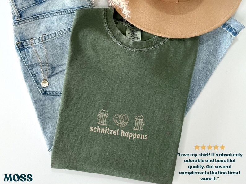 May include: An olive green t-shirt with the phrase "schnitzel happens" printed in white, along with beer mugs and a pretzel graphic. The shirt is laid on top of blue jeans and a tan hat.