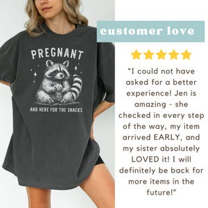 May include: A dark gray t-shirt with a graphic of a raccoon holding a jar of cookies. The text "PREGNANT AND HERE FOR THE SNACKS" is printed on the shirt.