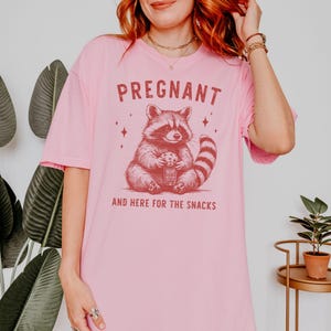 May include: Pink t-shirt with a graphic of a raccoon holding a cookie and the text "PREGNANT AND HERE FOR THE SNACKS".