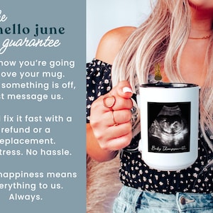 May include: White ceramic mug with a black rim, featuring a black and white ultrasound image and the text "Baby Thompson". The mug is held by a person. The mug is part of the "hello june guarantee".