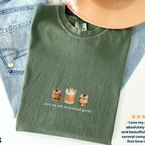Może przedstawiać: Oliwkowo-zielona koszulka z akwarelową ilustracją trzech koktajli i napisem "just an old fashioned girlie". Koszulka leży na niebieskich dżinsach i beżowym kapeluszu.
