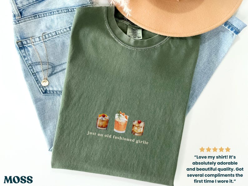 Może przedstawiać: Oliwkowo-zielona koszulka z akwarelową ilustracją trzech koktajli i napisem "just an old fashioned girlie". Koszulka leży na niebieskich dżinsach i beżowym kapeluszu.