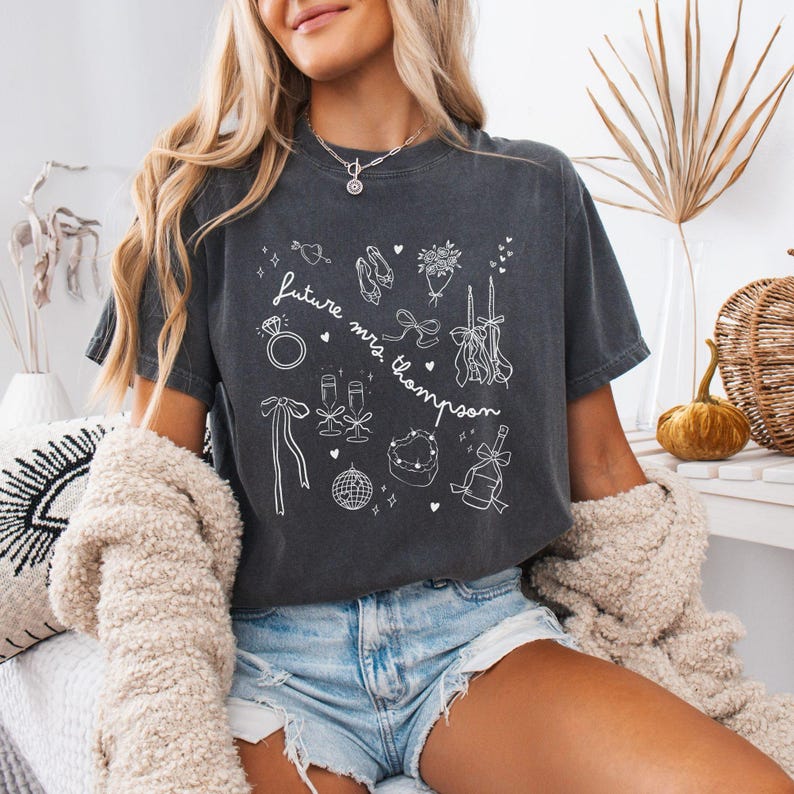 Puede incluir: Camiseta gris oscuro con ilustraciones en blanco de elementos relacionados con bodas, como un anillo, zapatos, copas de champ&aacute;n y un pastel en forma de coraz&oacute;n. El texto "future mrs. Thompson" tambi&eacute;n est&aacute; en la camiseta.
