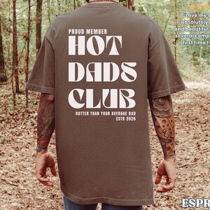 May include: Espresso-colored t-shirt with the text "HOT DADS CLUB" in white, along with "PROUD MEMBER" and "HOTTER THAN YOUR AVERAGE DAD ESTD 2026." The shirt is worn by a person in a forest setting.