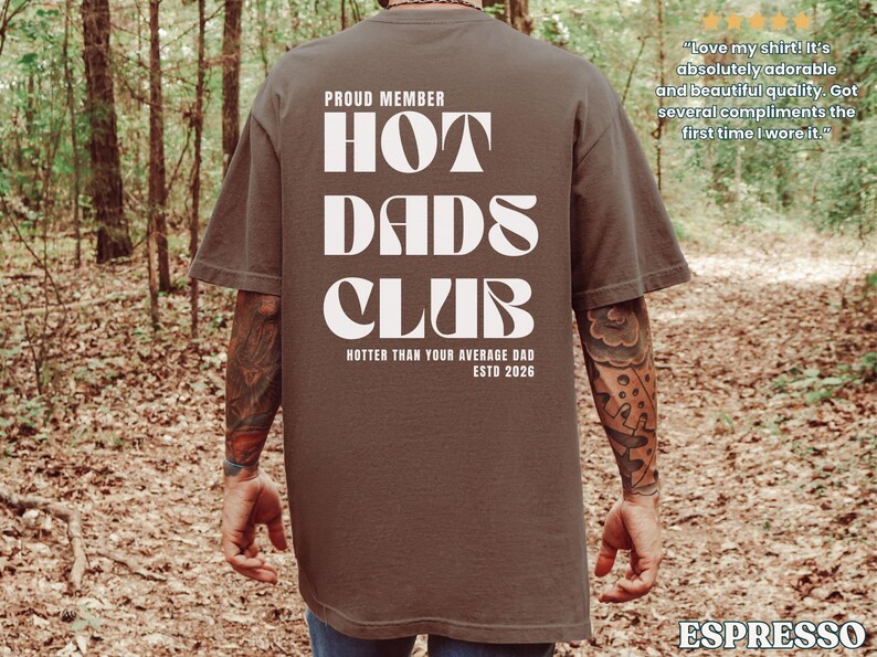 May include: Espresso-colored t-shirt with the text "HOT DADS CLUB" in white, along with "PROUD MEMBER" and "HOTTER THAN YOUR AVERAGE DAD ESTD 2026." The shirt is worn by a person in a forest setting.