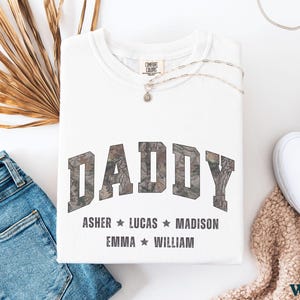 K&ouml;nnte beinhalten: Wei&szlig;es Sweatshirt mit dem Wort "DADDY" im Camouflage-Print. Darunter stehen die Namen Asher, Lucas, Madison, Emma und William. Das Sweatshirt liegt neben einer blauen Jeans, wei&szlig;en Turnschuhen und einer Halskette.