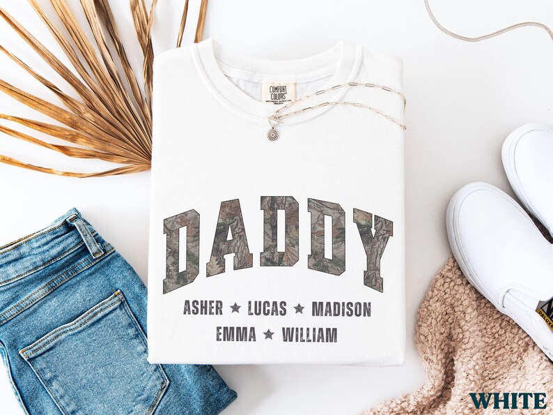 K&ouml;nnte beinhalten: Wei&szlig;es Sweatshirt mit dem Wort "DADDY" im Camouflage-Print. Darunter stehen die Namen Asher, Lucas, Madison, Emma und William. Das Sweatshirt liegt neben einer blauen Jeans, wei&szlig;en Turnschuhen und einer Halskette.