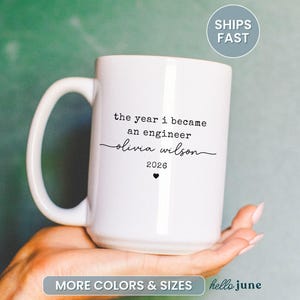 May include: A white ceramic mug with a large handle. The mug has the text "the year i became an engineer olivia wilson 2026" printed in black. A small black heart is below the year. The mug is held in a hand.