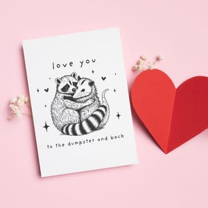 May include: A white card with a black and white illustration of a raccoon and opossum hugging. The card says "love you to the dumpster and back".