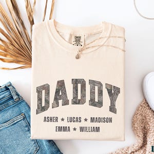 K&ouml;nnte beinhalten: Cremefarbenes T-Shirt mit dem Wort "DADDY" in einem Camouflage-Design. Darunter stehen die Namen Asher, Lucas, Madison, Emma und William. Das Shirt ist gefaltet mit blauer Jeans, wei&szlig;en Schuhen und einer Halskette.