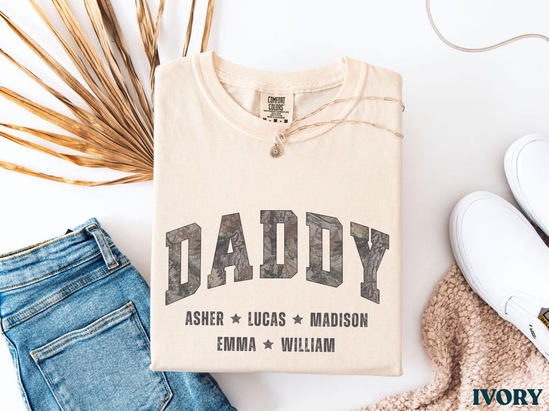 K&ouml;nnte beinhalten: Cremefarbenes T-Shirt mit dem Wort "DADDY" in einem Camouflage-Design. Darunter stehen die Namen Asher, Lucas, Madison, Emma und William. Das Shirt ist gefaltet mit blauer Jeans, wei&szlig;en Schuhen und einer Halskette.