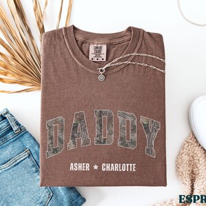 K&ouml;nnte beinhalten: Espressofarbenes T-Shirt mit dem Wort "DADDY" in einem Camouflage-Design. Die Namen "Asher" und "Charlotte" sind darunter aufgedruckt. Eine silberne Halskette und wei&szlig;e Turnschuhe sind ebenfalls zu sehen. Das Shirt liegt gefaltet auf einer wei&szlig;en Oberfl&auml;che.