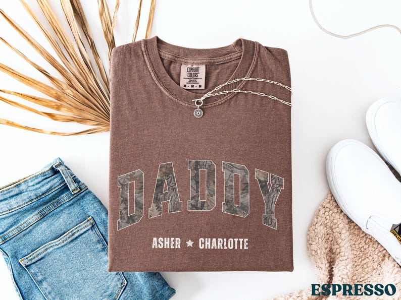 K&ouml;nnte beinhalten: Espressofarbenes T-Shirt mit dem Wort "DADDY" in einem Camouflage-Design. Die Namen "Asher" und "Charlotte" sind darunter aufgedruckt. Eine silberne Halskette und wei&szlig;e Turnschuhe sind ebenfalls zu sehen. Das Shirt liegt gefaltet auf einer wei&szlig;en Oberfl&auml;che.