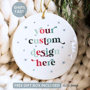 May include: A white ceramic ornament with the text "your custom design here" in various colors and snowflake accents. The ornament has a gold string for hanging and is set against a cozy, textured background.