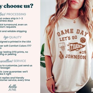 May include: Cream-coloured t-shirt featuring a brown graphic that says "GAME DAY LET'S GO TIGERS #25 JOHNSON" with a basketball, pennant, and basketball hoop design. The shirt is made of soft Comfort Colors material.