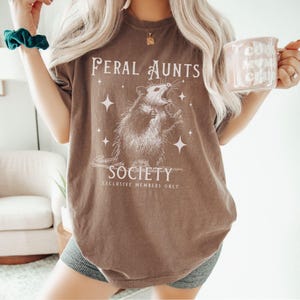 May include: A brown t-shirt with a graphic of a possum and the text "Feral Aunts Society Exclusive Members Only".