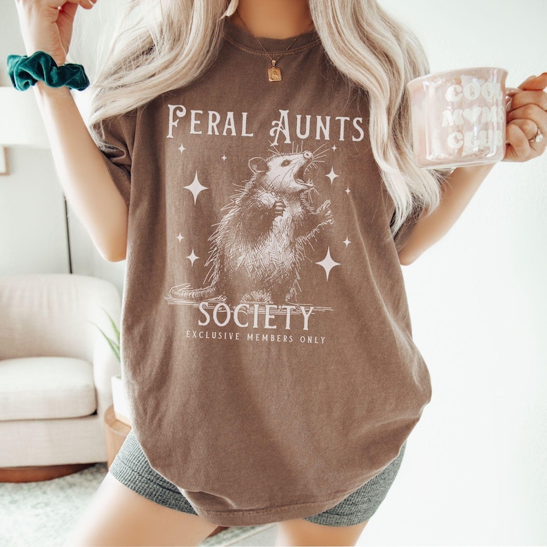 May include: A brown t-shirt with a graphic of a possum and the text "Feral Aunts Society Exclusive Members Only".