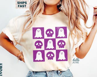 May include: Cream-coloured t-shirt with a grid of purple squares, each containing a ghost or skull design. The ghosts have smiling faces. The text "hello JUNE" is on the lower right. The shirt is a casual style.