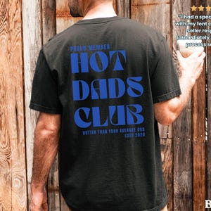 May include: Black t-shirt with the text "HOT DADS CLUB" in blue, with "PROUD MEMBER" above and "HOTTER THAN YOUR AVERAGE DAD ESTD 2026" below. The shirt is a casual style.