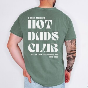 May include: Moss green t-shirt with the text "PROUD MEMBER HOT DADS CLUB" in white, along with "HOTTER THAN YOUR AVERAGE DAD ESTD 2026". The shirt is a relaxed fit.