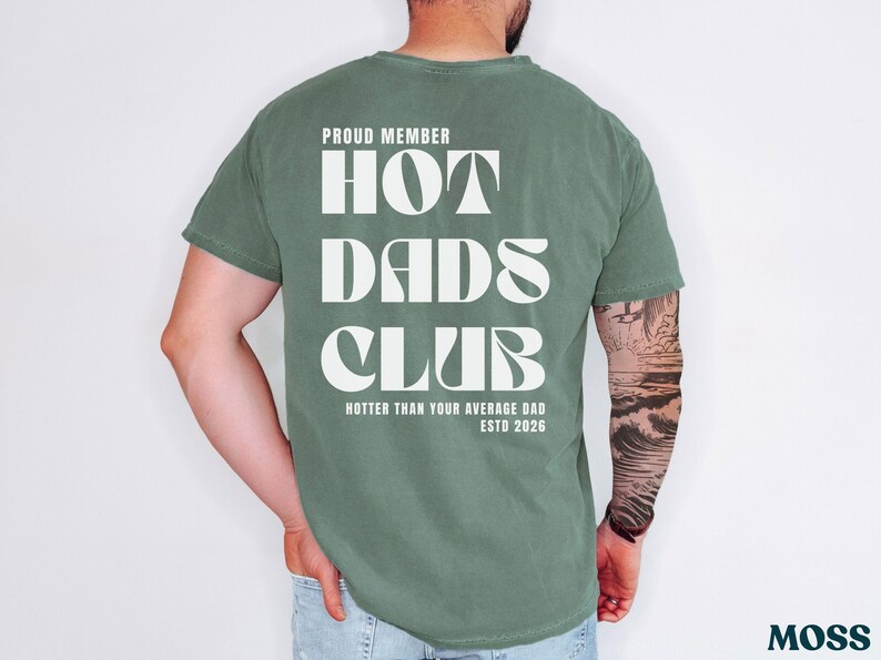 May include: Moss green t-shirt with the text "PROUD MEMBER HOT DADS CLUB" in white, along with "HOTTER THAN YOUR AVERAGE DAD ESTD 2026". The shirt is a relaxed fit.