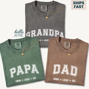 May include: Three t-shirts in gray, green, and brown, each with a different word in white block letters: "GRANDPA", "PAPA", and "DAD". Each shirt has a gold star necklace. The names Emma, Lucas, and Ava are printed below each word.