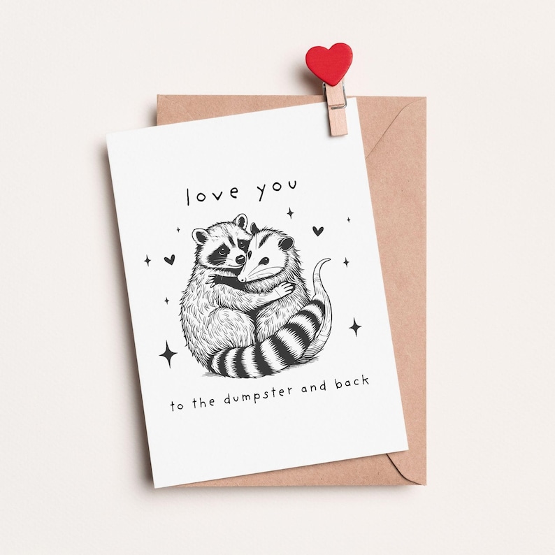 May include: A white card with a black and white illustration of a raccoon and opossum hugging. The card says "love you to the dumpster and back".