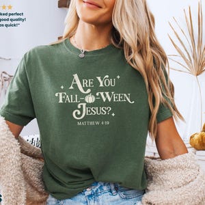 May include: Green t-shirt with the text "Are You Fall-Ween Jesus?" and "Matthew 4:19" in white. The shirt has a relaxed fit and is made of a soft material. The design includes a pumpkin graphic. The shirt is part of a fall-themed collection.