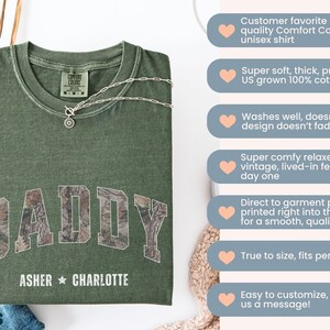 K&ouml;nnte beinhalten: Ein olivgr&uuml;nes Unisex-T-Shirt mit dem Wort "DADDY" in einem Camouflage-Muster auf der Brust. Darunter stehen die Namen "ASHER" und "CHARLOTTE". Eine silberne Halskette mit Anh&auml;nger liegt auf dem Shirt.