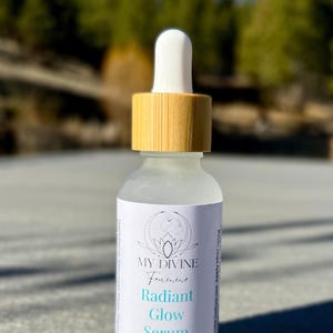 May include: A clear glass dropper bottle with a light wood-toned cap and a white dropper top. The label features the text MY DIVINE Feminine Radiant Glow Serum and 1 Fl oz, with a circular line art logo of a face above the product name.