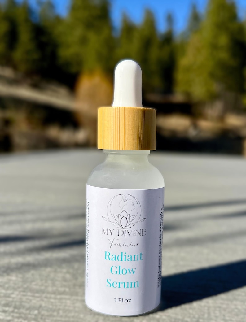 May include: A clear glass dropper bottle with a light wood-toned cap and a white dropper top. The label features the text MY DIVINE Feminine Radiant Glow Serum and 1 Fl oz, with a circular line art logo of a face above the product name.