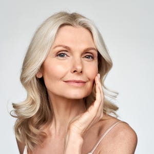 May include: A person with shoulder-length blonde hair and blue eyes, looking directly forward with a gentle smile. One hand is positioned near the jawline, showcasing a clear complexion. The individual wears a thin-strapped garment against a plain background.