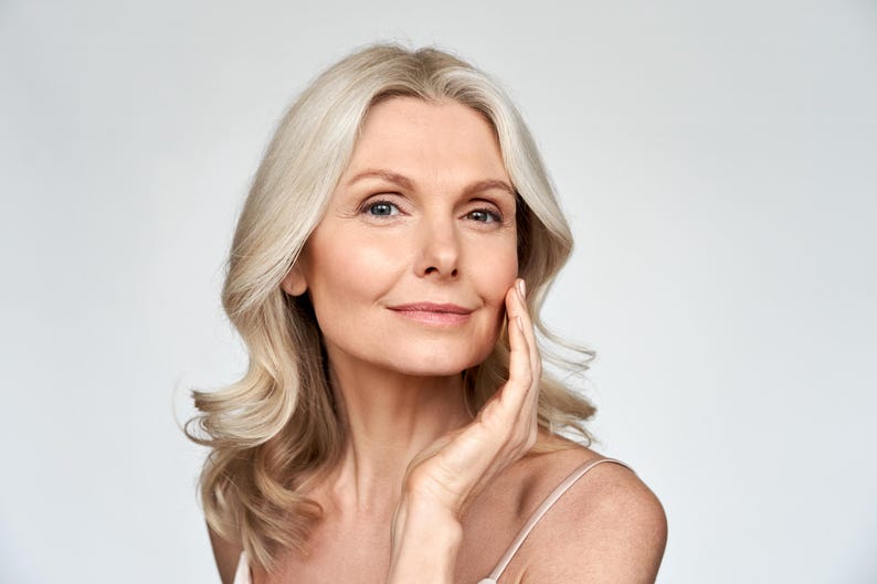 May include: A person with shoulder-length blonde hair and blue eyes, looking directly forward with a gentle smile. One hand is positioned near the jawline, showcasing a clear complexion. The individual wears a thin-strapped garment against a plain background.